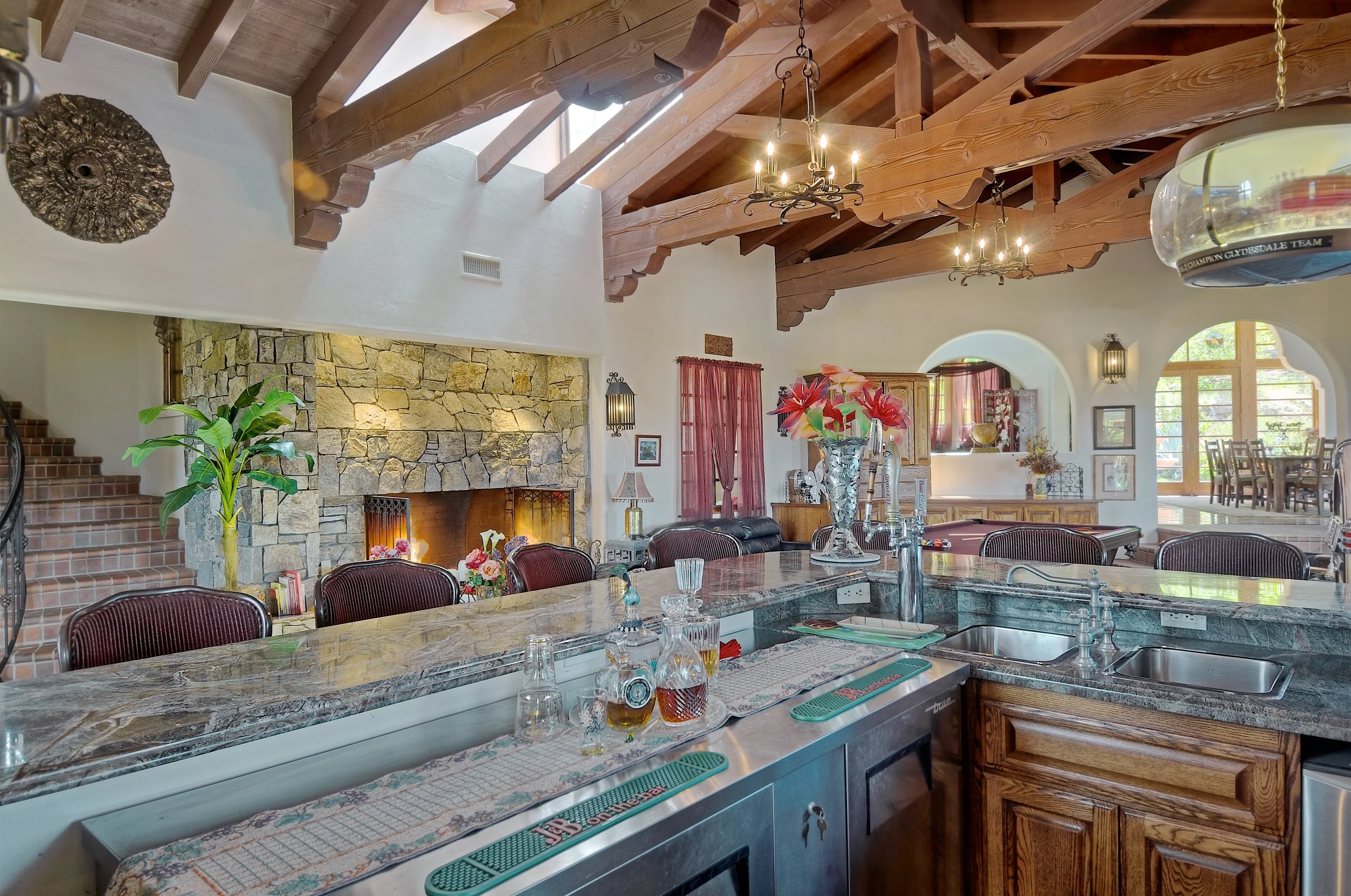 A spacious living room with a high wooden beam ceiling, a stone fireplace, a large table with chairs, and floral decorations.