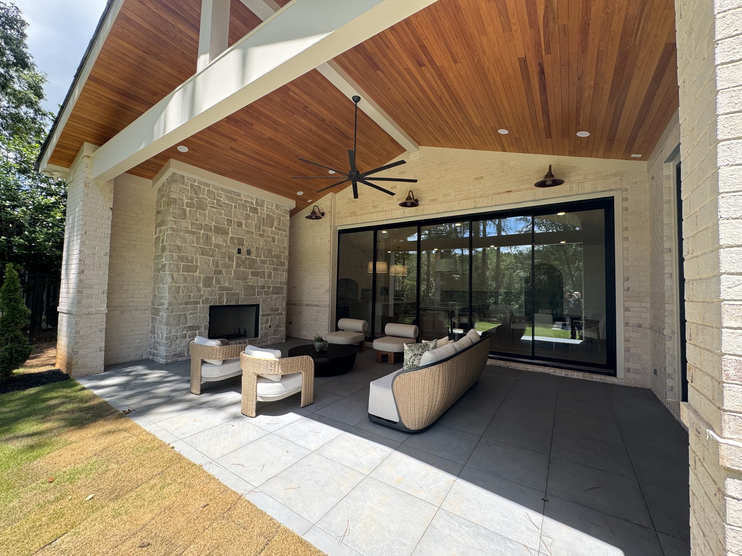 An outdoor patio area with wicker chairs, a cushioned sofa, and a coffee table, shaded by a wooden ceiling with a ceiling fan. The patio is adjacent to a house with sliding glass doors, brick walls, and hanging outdoor lamps. There is a grassy yard s