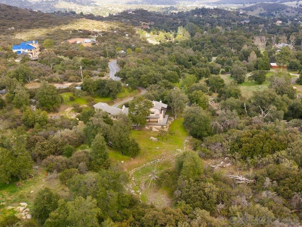 Aerial view of a house surrounded by trees and greenery, with a winding road nearby in a hilly landscape.