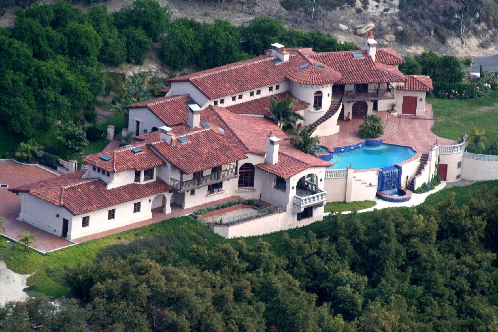 A large Mediterranean-style mansion with red tile roof, surrounded by greenery, featuring a kidney-shaped swimming pool with water features and a waterfall.