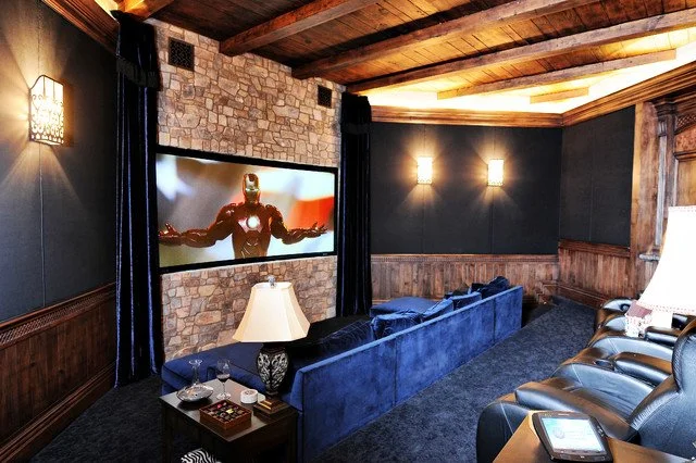Cozy home theater room with a large screen displaying Iron Man, dark walls, wooden ceiling, and blue and leather seats.