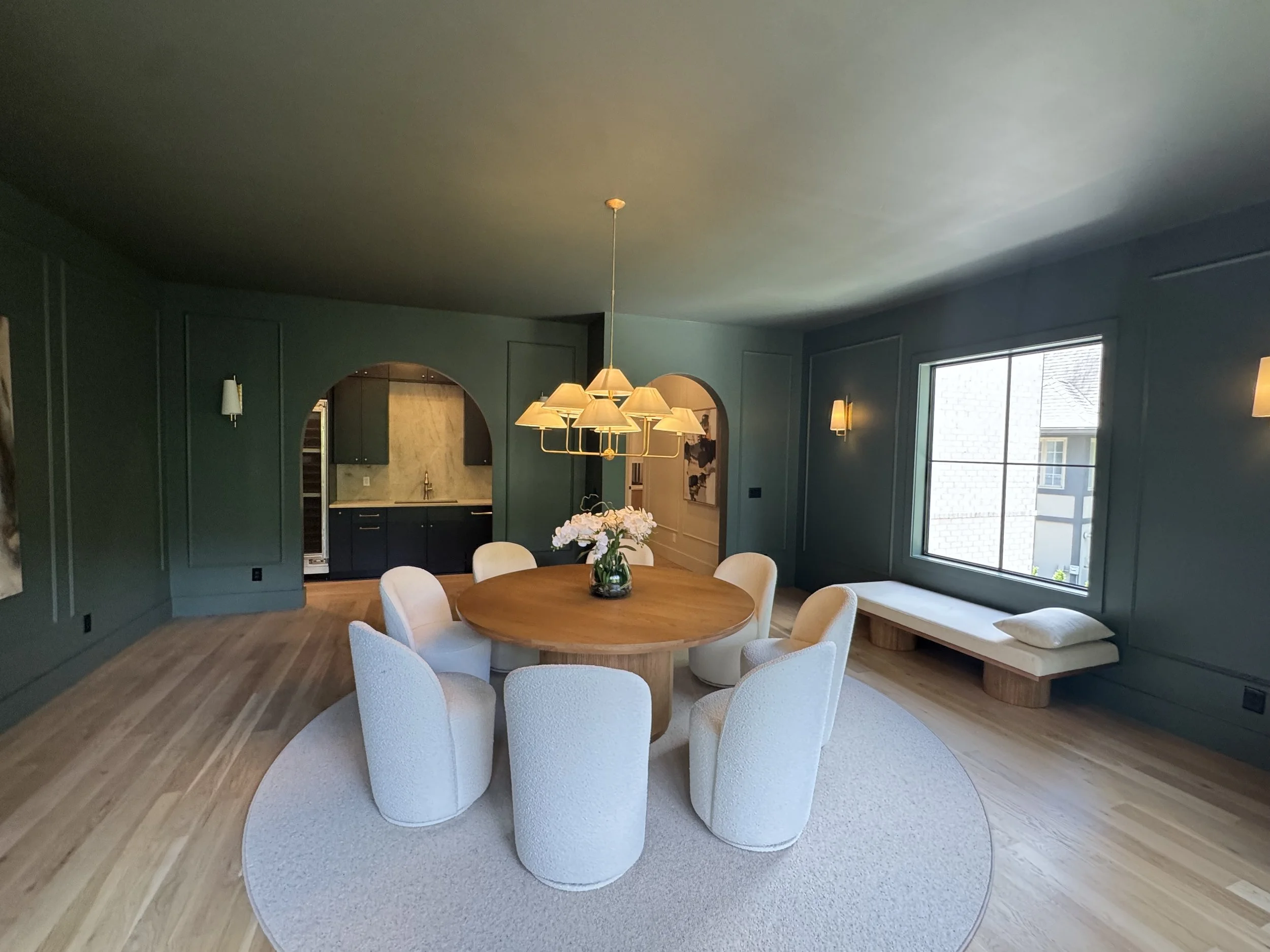 A dining room with a round wooden table surrounded by eight white upholstered chairs. A vase with white flowers sits on the table. The room has dark green walls, large windows, and a modern chandelier with multiple lamps.