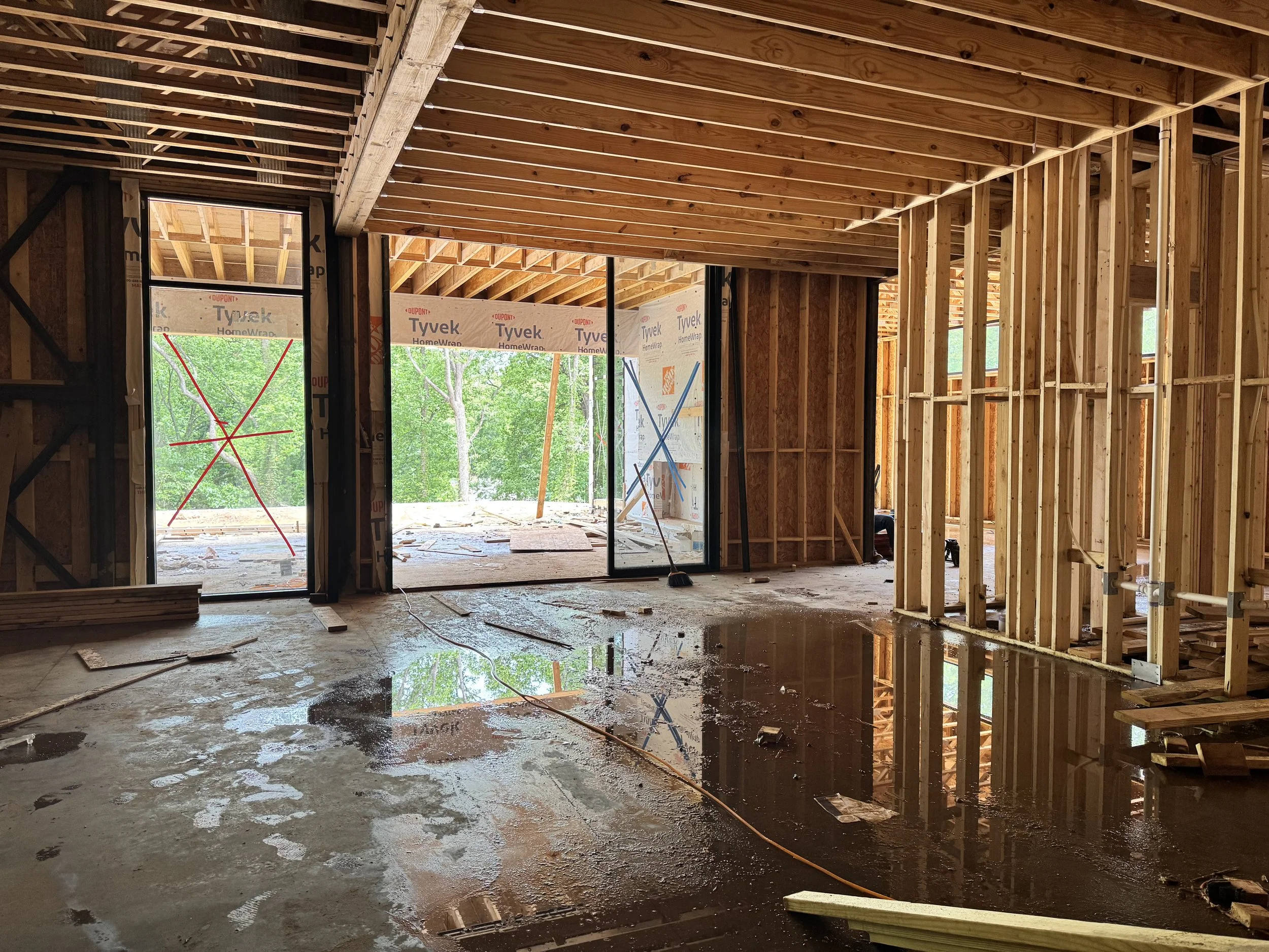 Interior view of a house under construction with exposed wooden framing, unfinished walls, and a wet concrete floor with puddles, visible through large open window and door spaces.