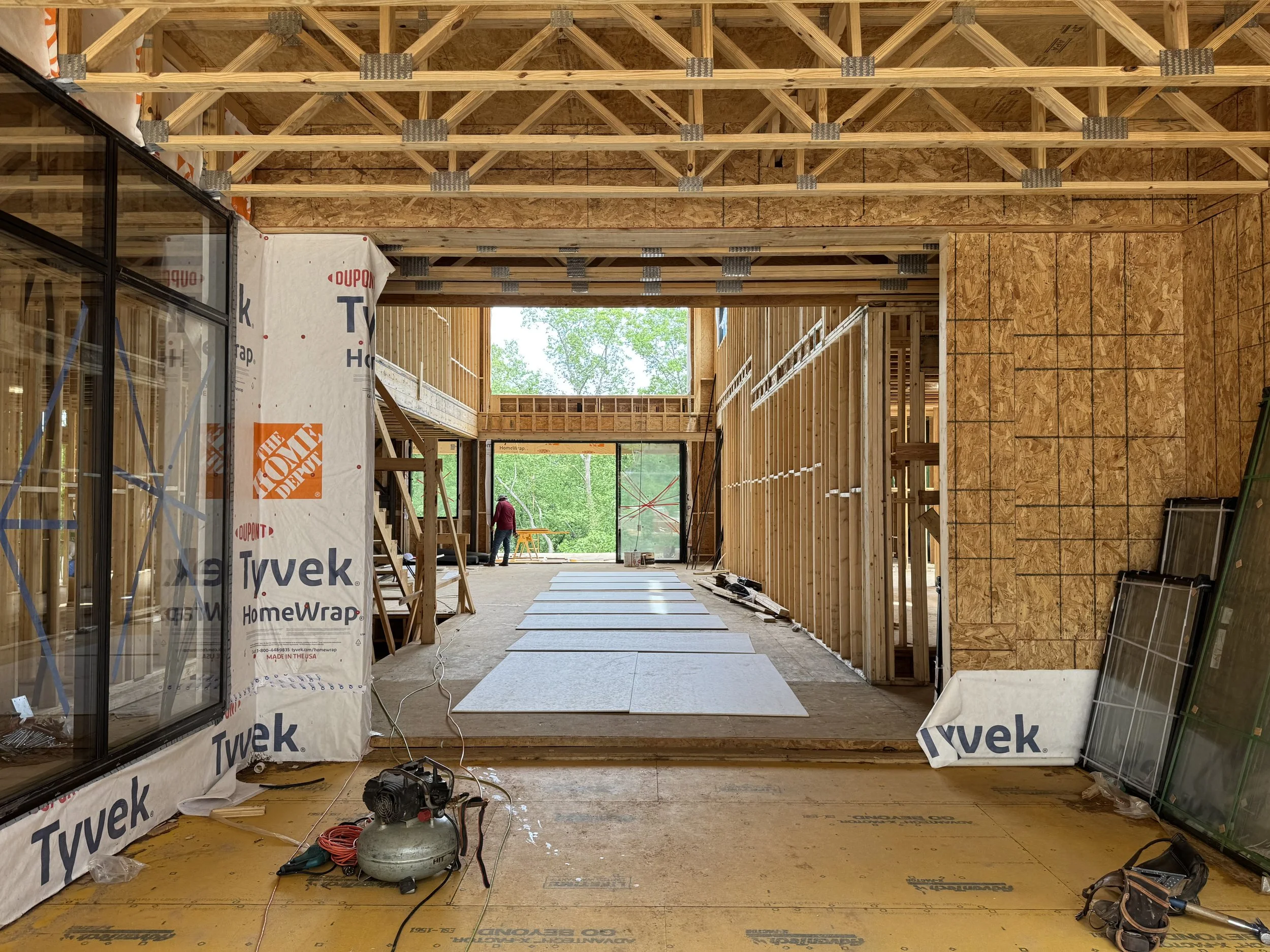 Interior view of a building under construction, showing exposed wooden framing, drywall, and flooring. Construction materials and tools are scattered around, and a person is working in the background near a tree outside.