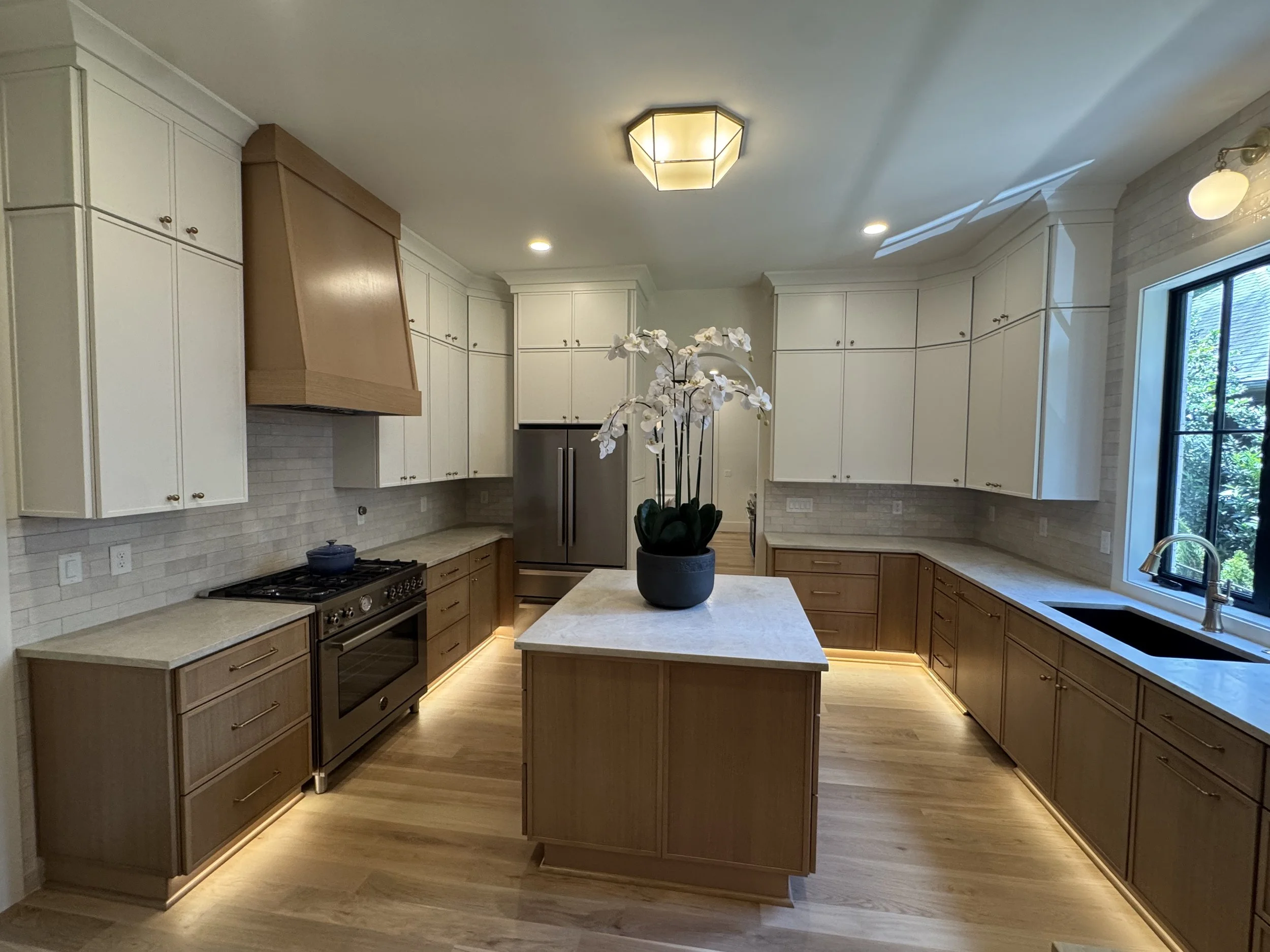 Kitchen with white cabinets, a kitchen island with a black potted plant, a stainless steel refrigerator, a stove, and a window with natural light and greenery outside.