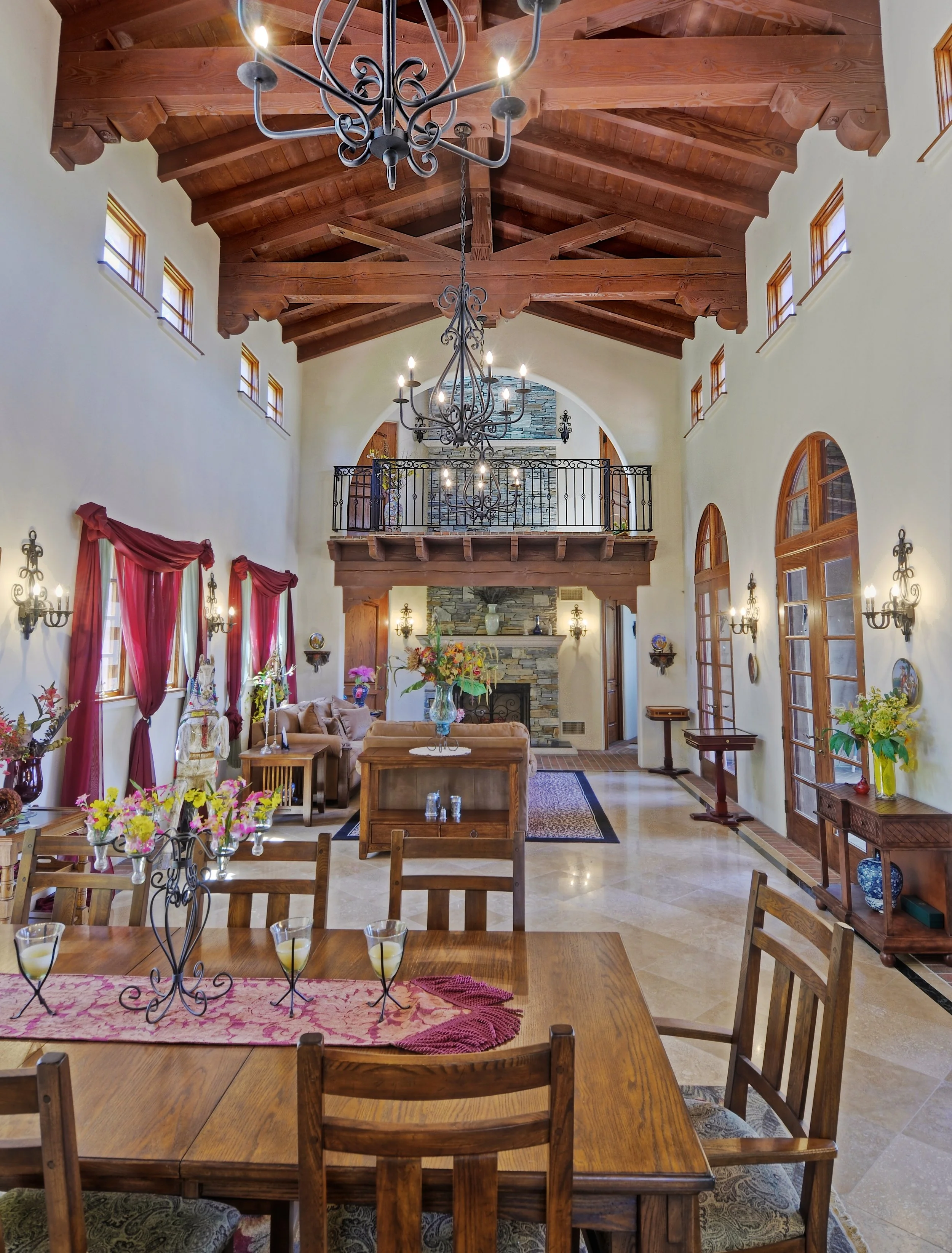 Interior view of a spacious living room with a high wooden vaulted ceiling, chandeliers, a stone fireplace, and large windows with red curtains. There are tables with flowers and candles, and a balcony with a wrought-iron railing.