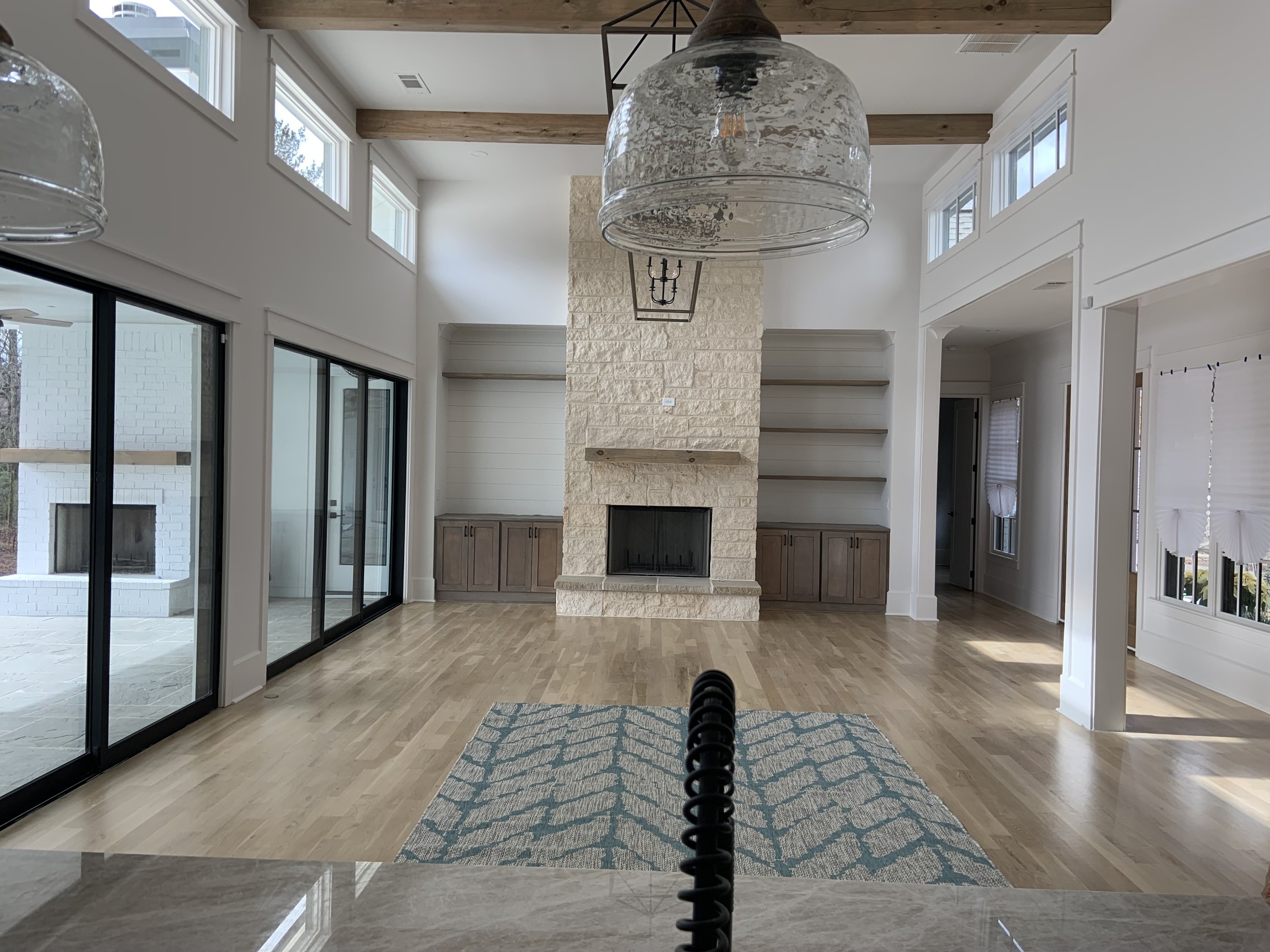 It is a spacious living room with a high ceiling, large windows, a stone fireplace, wooden beams on the ceiling, a patterned area rug, and sliding glass doors leading to an outdoor patio.