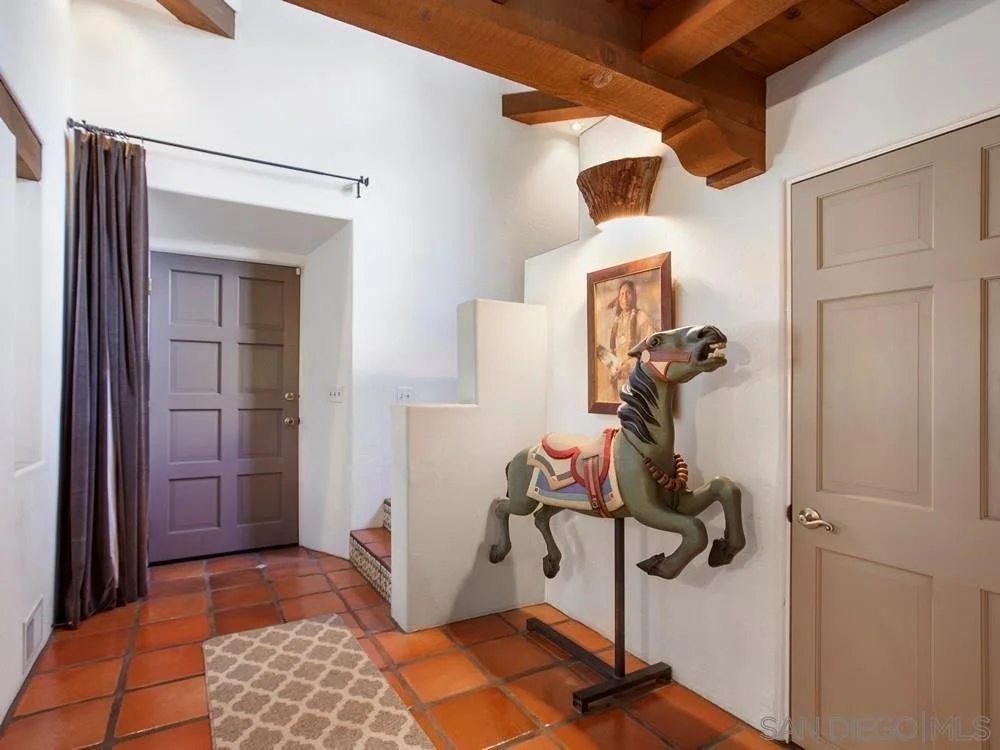 Entryway of a house with a brown front door, a gray interior door, terracotta tile flooring, a small beige patterned rug, and a decorative carousel horse sculpture next to a framed portrait on the white wall. The ceiling has wooden beams.