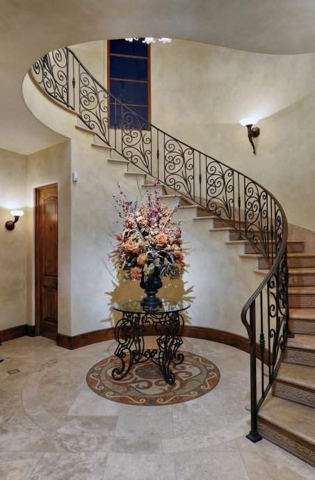 Elegant curved staircase with wrought iron railing, a flower arrangement on a decorative table, and wall-mounted light fixtures in a luxurious interior.