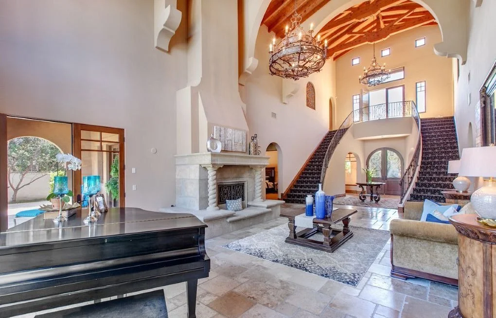 Spacious living room featuring a grand staircase, fireplace, large windows, and chandelier with elegant furniture and decor.