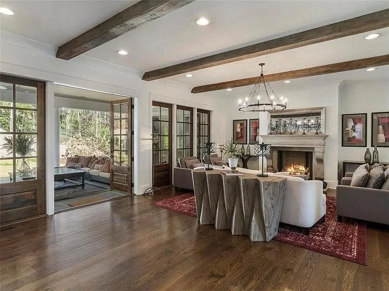 Spacious living room with a wood floor, a fireplace, a round chandelier, multiple grey sofas, a unique wooden dining table, and large windows showing a green outdoor area.