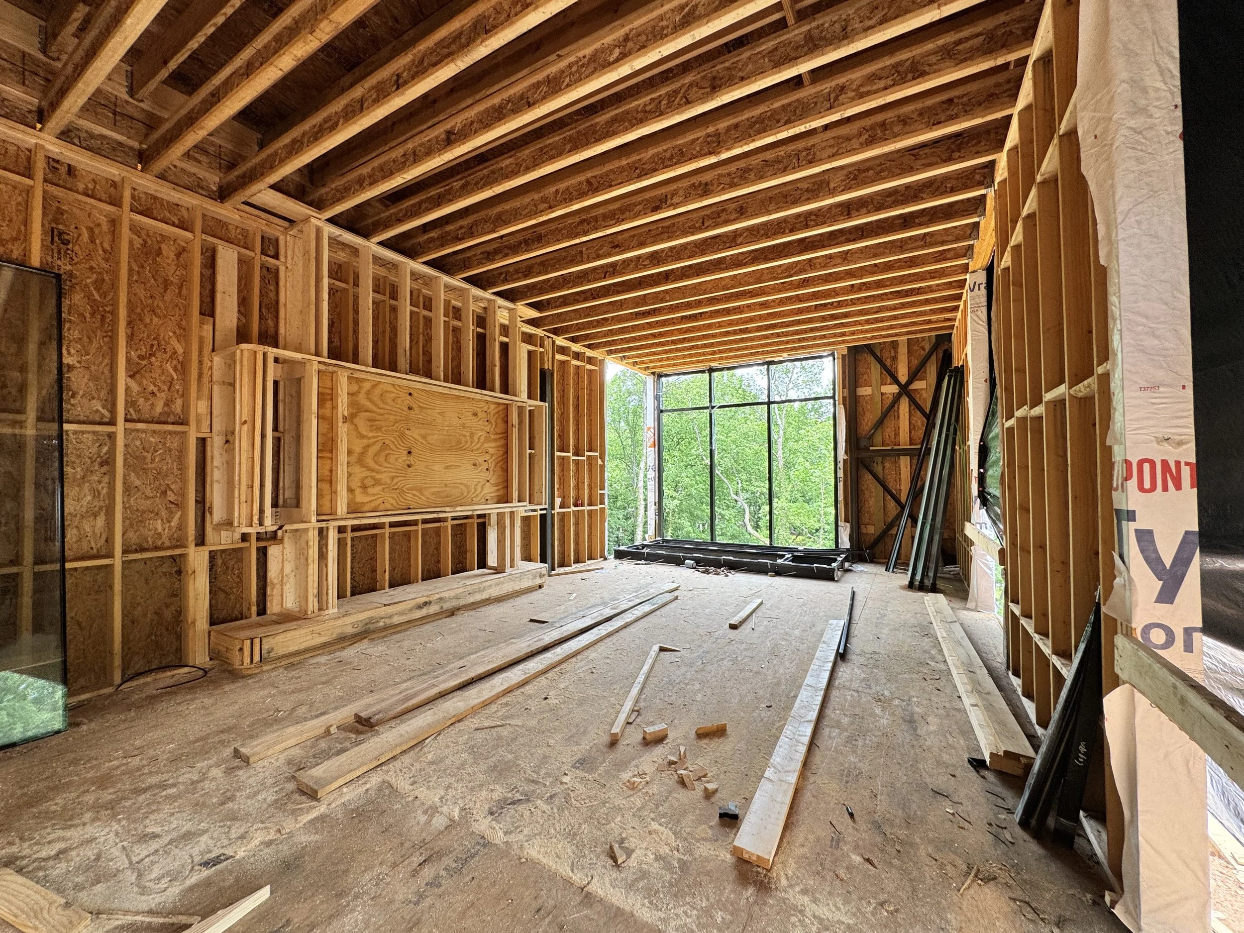 Interior of a house under construction with exposed wooden framing, unfinished walls, and a large window at the back showing green trees outside.