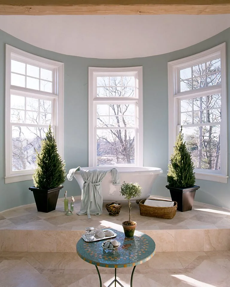 A bright bathroom with a white clawfoot bathtub placed on a raised beige tiled platform, flanked by three large windows with white frames, a small potted tree in front of the tub, and two potted plants with tall green foliage on either side. There is