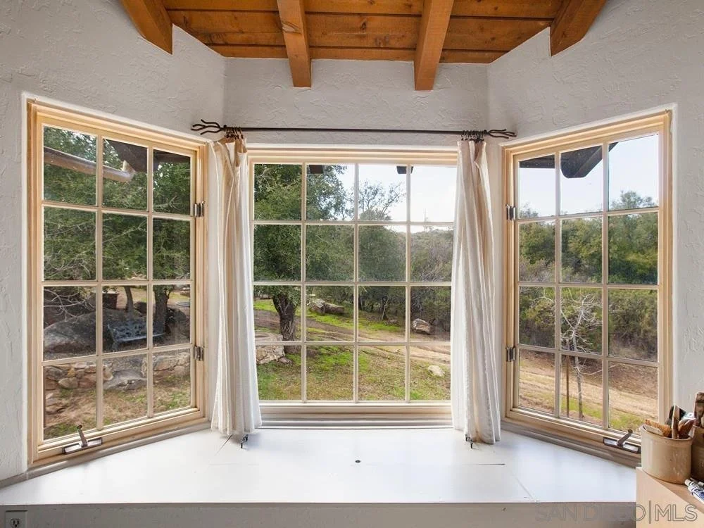 A three-pane window with white curtains, looking out onto a natural landscape with trees, rocks, and dirt path under a wooden ceiling.