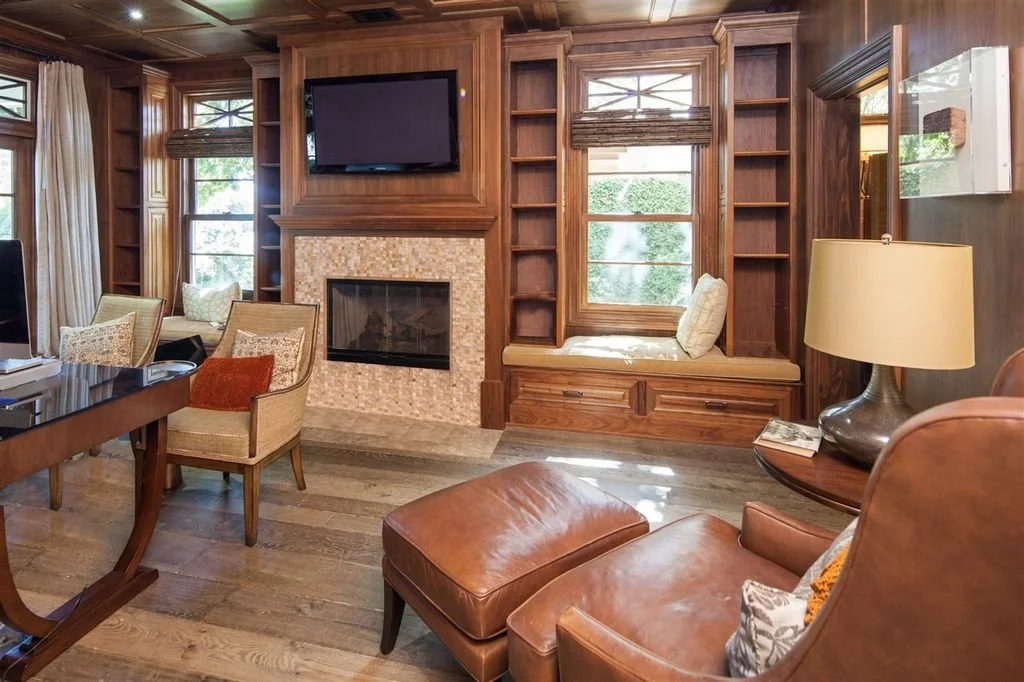 Living room with wooden bookshelves and built-in window seat, fireplace with mosaic tile surround, flat-screen TV above fireplace, leather armchair with matching ottoman, side table with lamp, and multiple windows with curtains