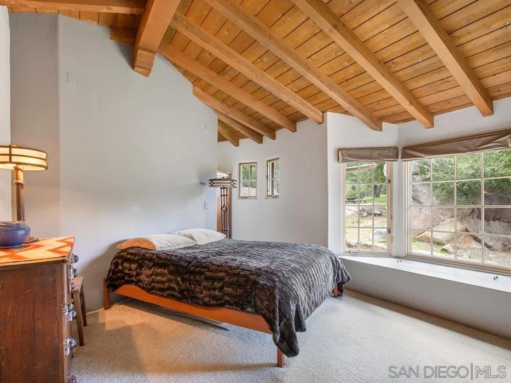 Bedroom with a sloped wooden ceiling, a large window with a view of outdoor rocks and greenery, a bed with dark bedding, a side table with a lamp, and a vintage wooden dresser.