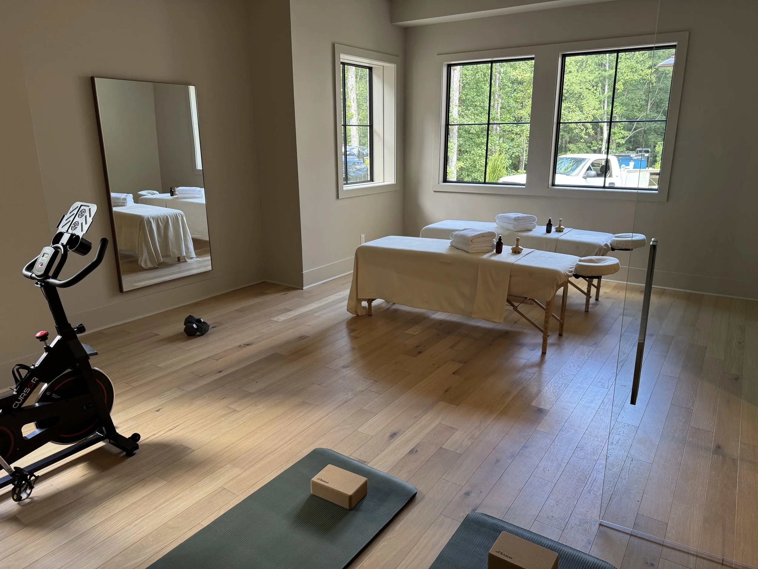 A room set up for a yoga or meditation class with two mats, yoga blocks, and bolsters, next to large windows with a view of trees outside, and a massage table with towels and oils.