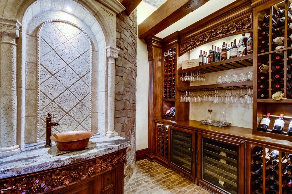 Wine storage room with wooden shelves filled with wine bottles, glassware, and a wine refrigerator, next to a stone and wood decorative fountain with a small basin.