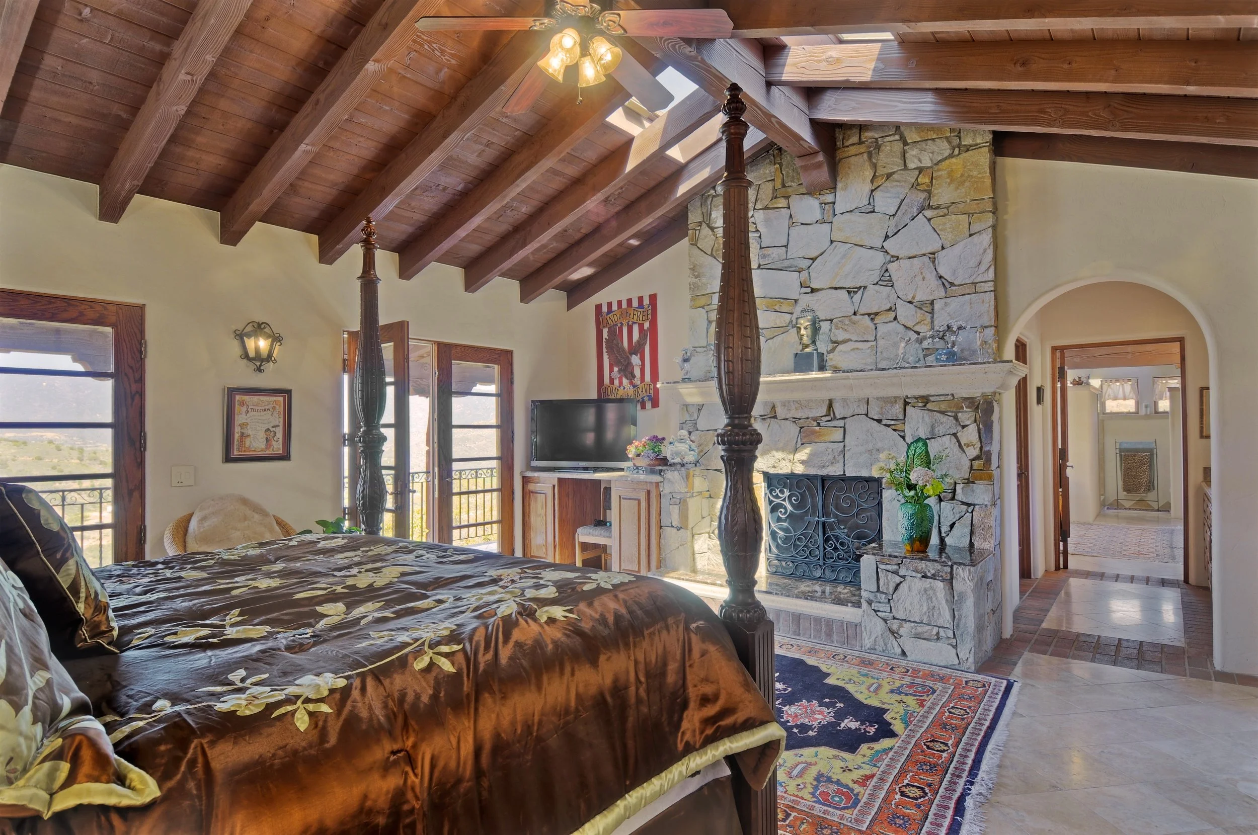 Interior of a rustic living room with a wooden vaulted ceiling, a stone fireplace, a ceiling fan with lights, a flat-screen TV, a floral bedspread, and a colorful area rug.