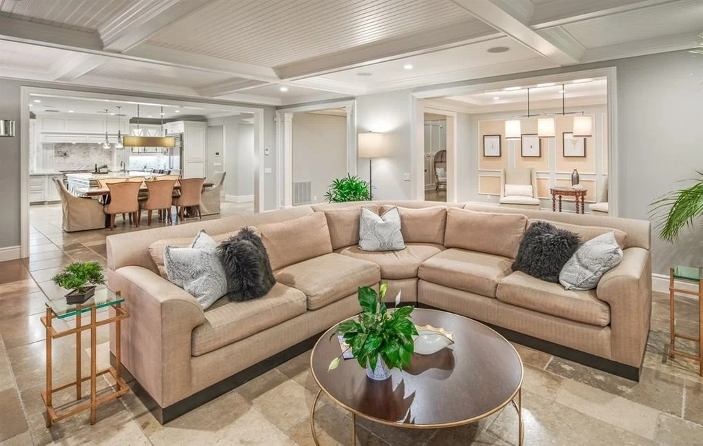 Spacious living room with beige sectional sofa, decorative pillows, round coffee table with green plant, flanking side tables with small plants, open to dining and kitchen areas, modern decor, bright lighting.