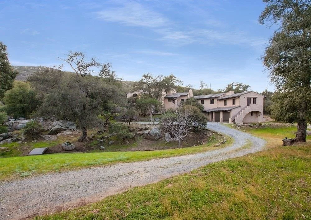 A house with adobe-style walls and a gravel driveway, surrounded by trees, rocks, and grass under a partly cloudy sky.