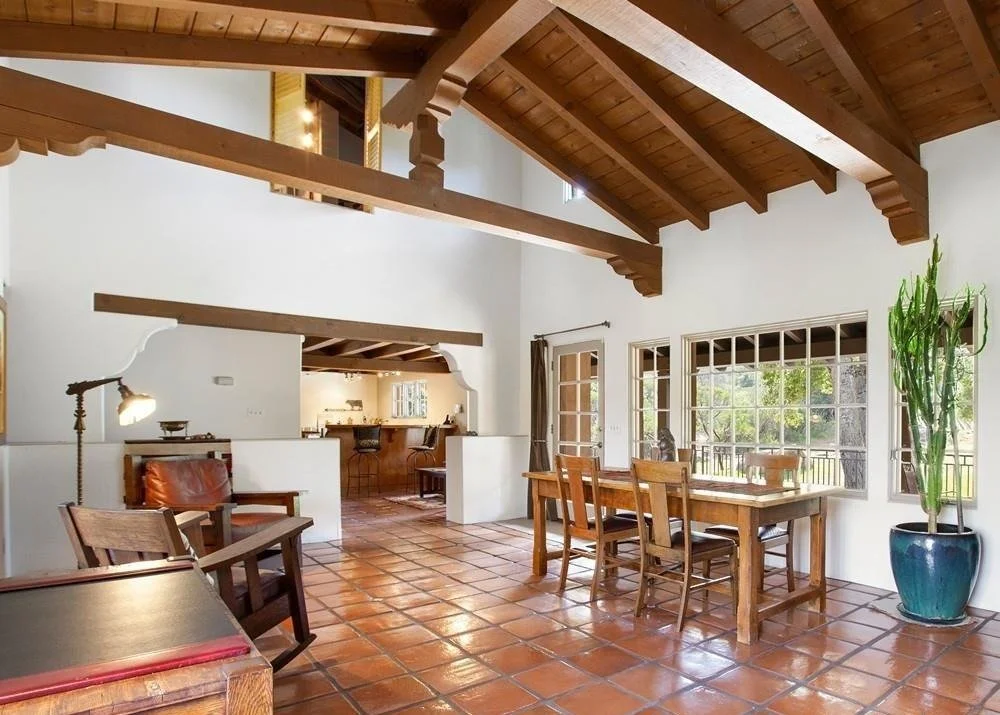 Living room with vaulted wooden ceiling, tiled floor, large windows, and wooden furniture including a dining table and chairs.