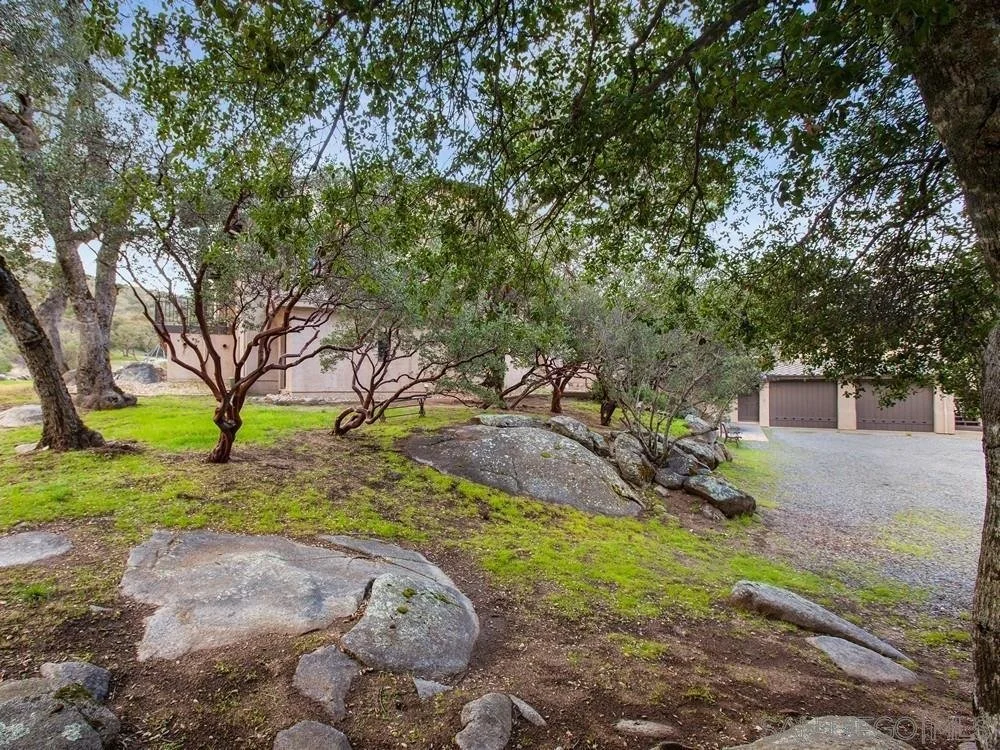 A landscaped yard with several trees, large rocks, and grasses, leading to a building with three garage doors.