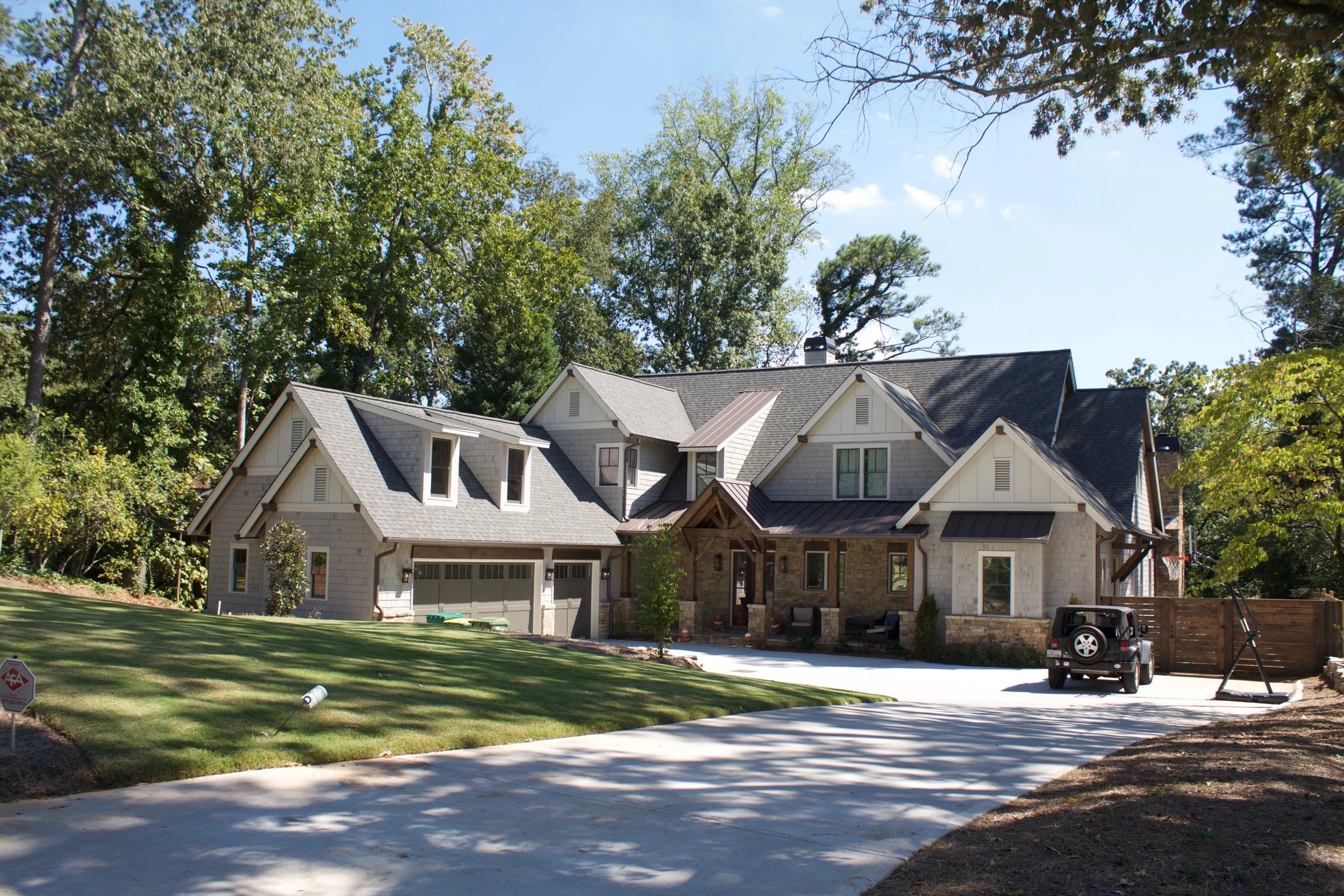 A large house with multiple peaked roofs surrounded by tall green trees on a sunny day, with a clear blue sky.