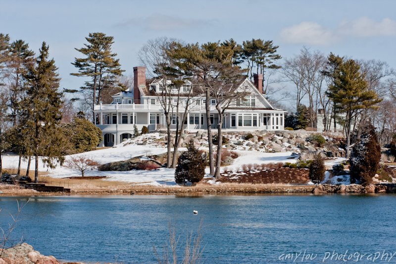 Large lakeside house with multiple stories, surrounded by snow, trees, and rocks, under a partly cloudy sky.