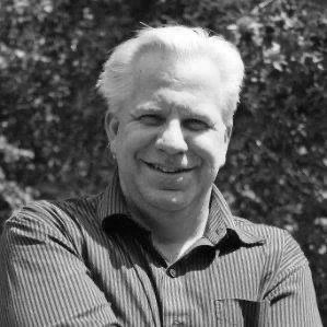 Black and white photo of a smiling man with white hair wearing a striped shirt, outdoors with trees in the background.