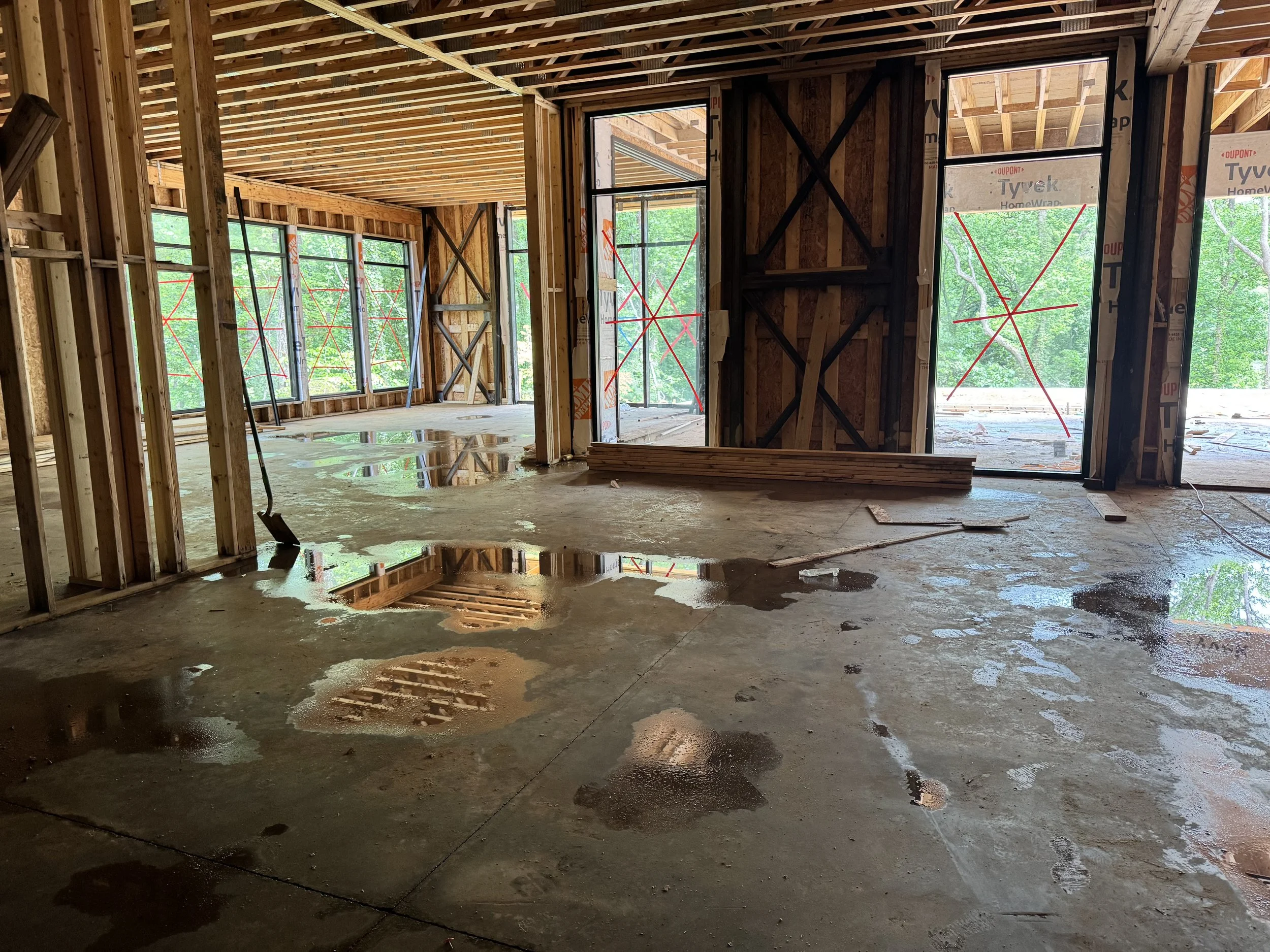 The interior of a house under construction showing exposed wooden framing, large windows, and a concrete floor with water puddles.