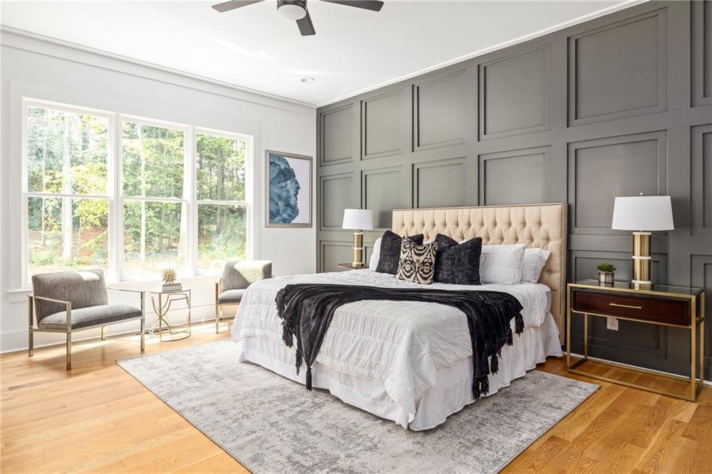 Modern bedroom with large window, gray paneled accent wall, beige upholstered bed with black and beige pillows, white bedding, two matching nightstands with white lamps, two gray chairs by the window, wooden floor, and a light area rug.