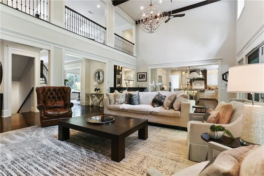 Spacious living room with high ceiling, white walls, large white sofa with pillows, a leather armchair, a black coffee table, and a beige armchair with a lamp, open to the kitchen area.