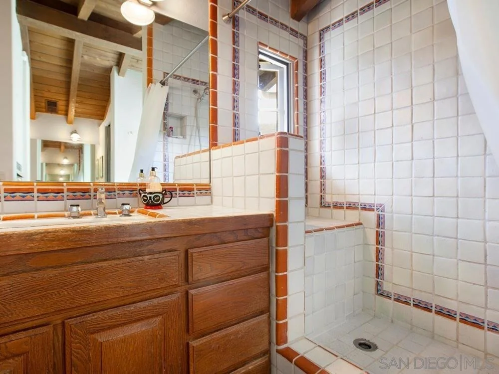Bathroom with wooden vanity, tile walls, a shower area with a small window, and a mirror reflecting the hallway.
