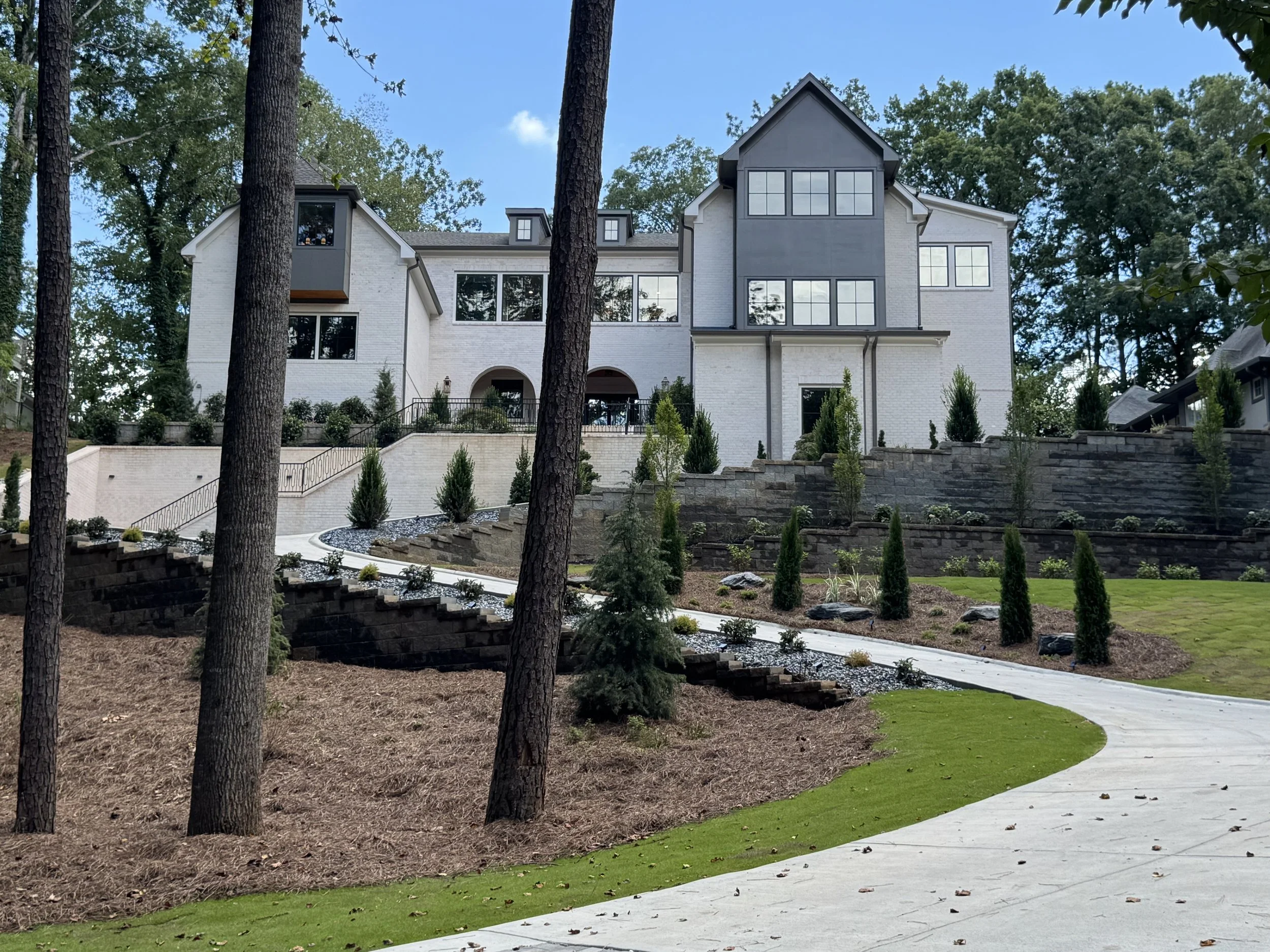 A modern white brick house with large windows is nestled among tall trees. The house features multiple levels with a prominent central gable and additional gable dormers. The front yard has a landscaped garden with small trees and shrubs, terraced re