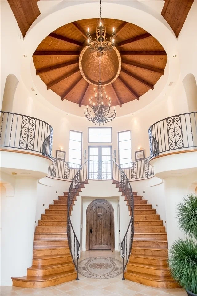 Elegant spiral staircase with wooden steps and wrought iron railings inside a bright, airy entrance hall with large windows and a wooden ceiling.
