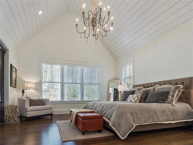 A bedroom with a large bed that has multiple pillows, a tufted headboard, and a patterned blanket. There is a red ottoman at the foot of the bed, a white armchair with a pillow and a lamp on a side table nearby. The room has large windows with blinds