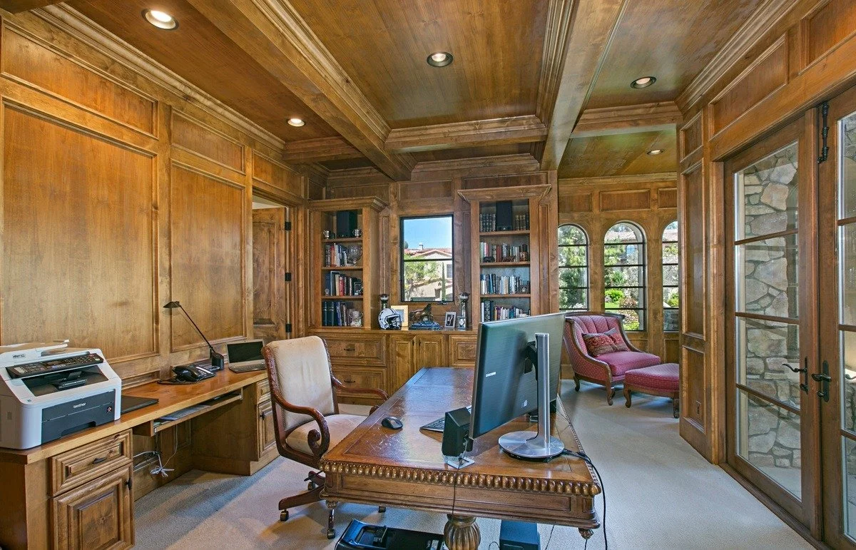 A home office with wood-paneled walls, a large wooden desk with a computer, and a bookshelf filled with books. There is a pink upholstered chair with an ottoman near arched windows showing a view of a backyard with trees. A printer is on a side desk,