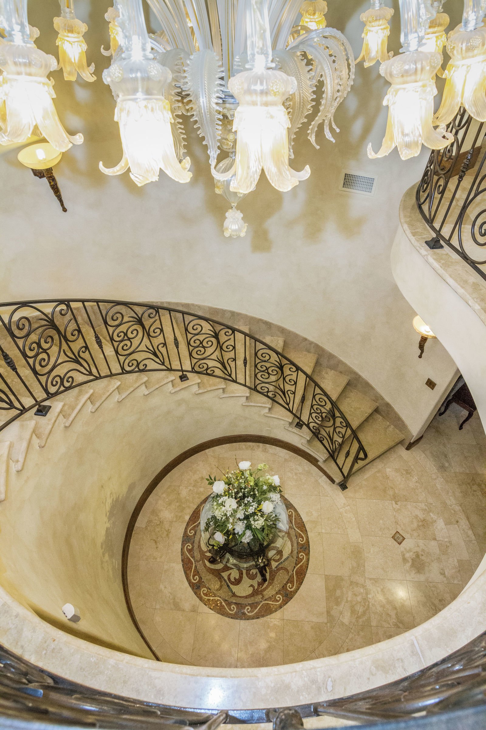 A grand chandelier hanging from a ceiling at the top of a curved staircase with wrought iron railing, overlooking a round marble floor with a floral arrangement in the center.
