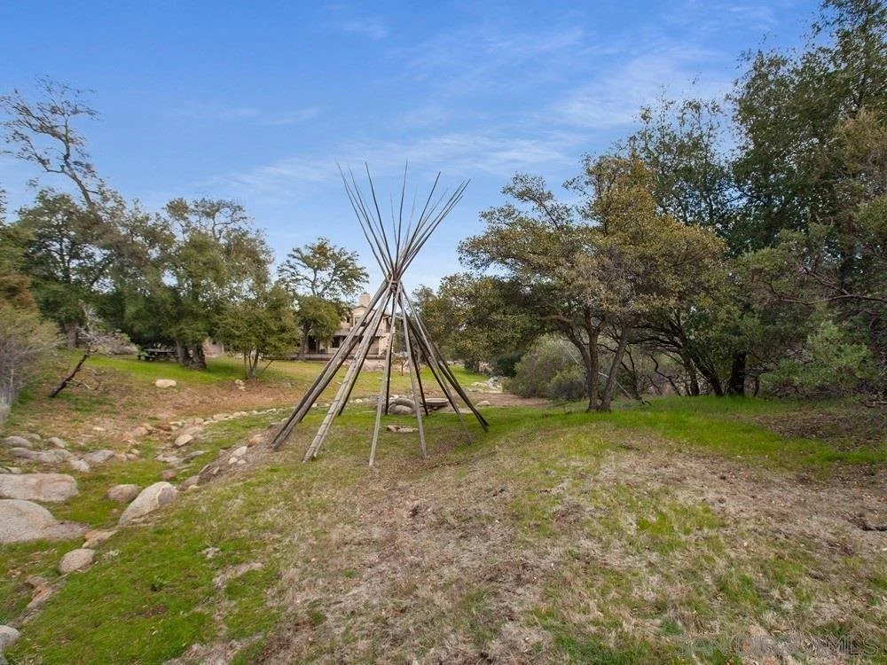 A traditional Apache tipi made of long wooden poles standing in a grassy, rocky outdoor area surrounded by trees and shrubs.