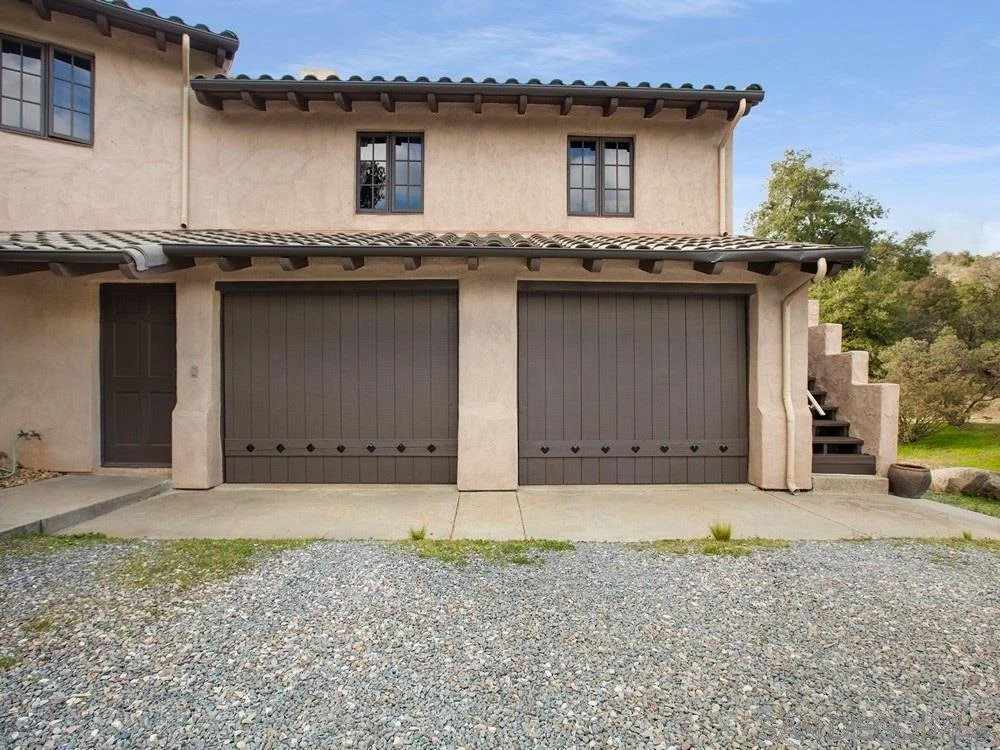 Two brown garage doors in a stucco house with a small window above each garage, a side door to the left, a staircase on the right, and a gravel driveway in front.