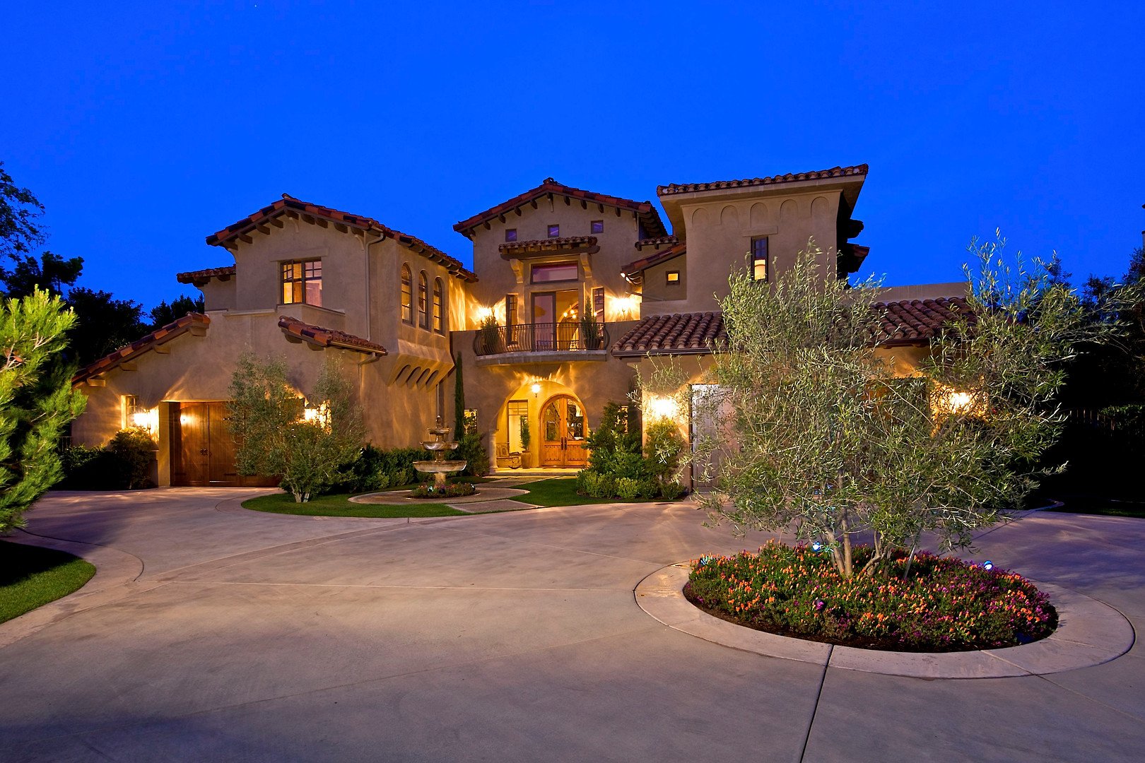 Large, luxurious house with warm exterior lighting, Spanish-style roof tiles, and a curved driveway, at dusk with a deep blue sky. Front yard features a small tree surrounded by colorful flowers, and a fountain at the entrance.