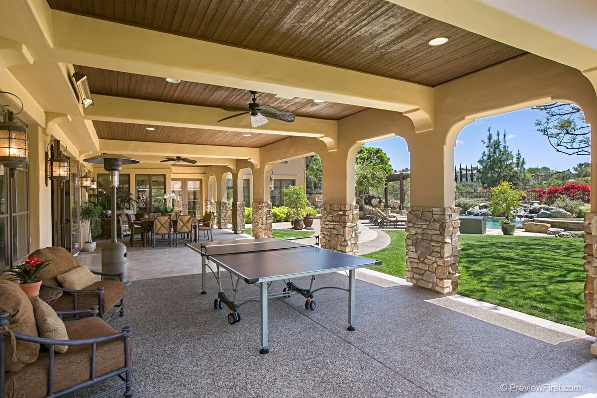 Covered outdoor patio with ping pong table, outdoor seating, ceiling fans, and a view of a landscaped yard with a pool and garden.