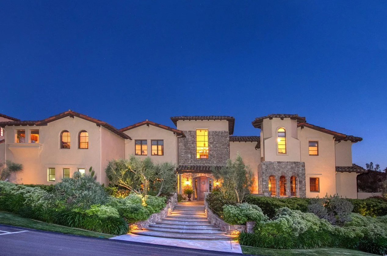 A large, luxurious house at dusk with warm interior lights, stone accents, multiple windows, surrounded by landscaped greenery and a pathway leading to the front door.