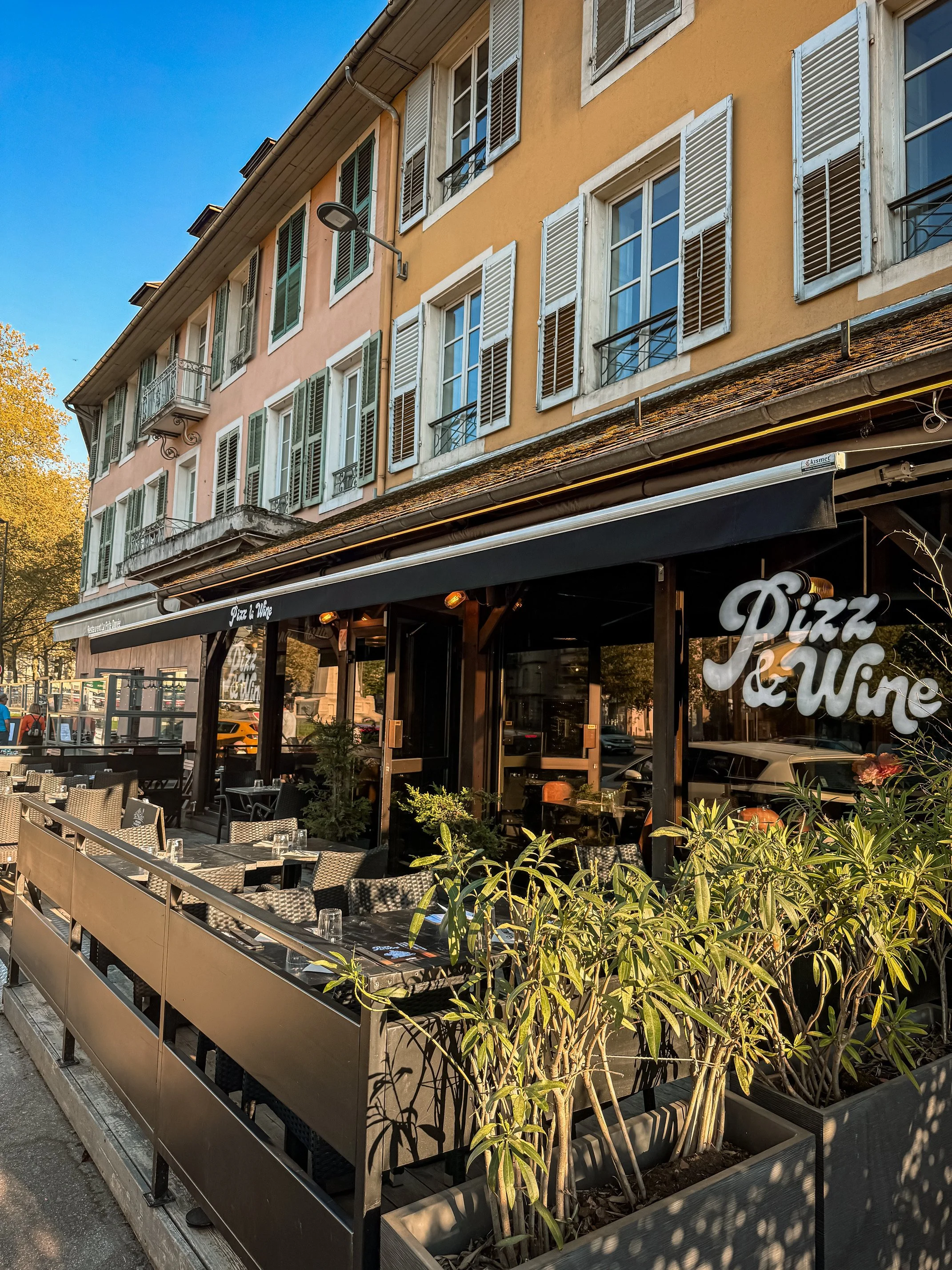 Terrasse de restaurant avec des tables, des chaises et des plantes, devant un bâtiment avec des fenêtres et des volets colorés. Signage disant 'Pizza & Wine'.