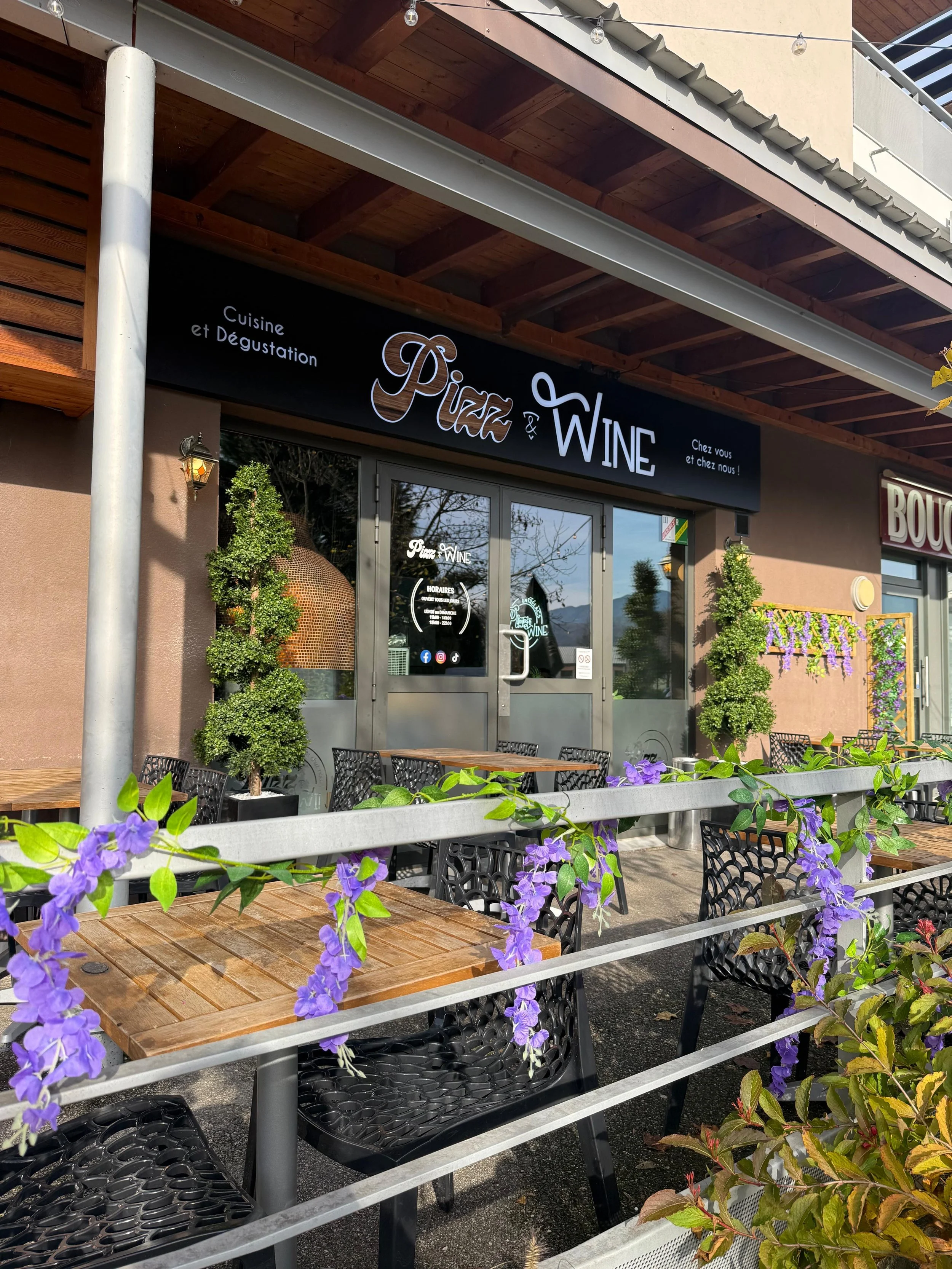 Façade d'une restaurant ou bar appelé Pizza & Wine avec un panneau noir, fenêtres, tables en bois et chaises en plastique noir, et des plantes vertes avec des fleurs violettes à l'avant.