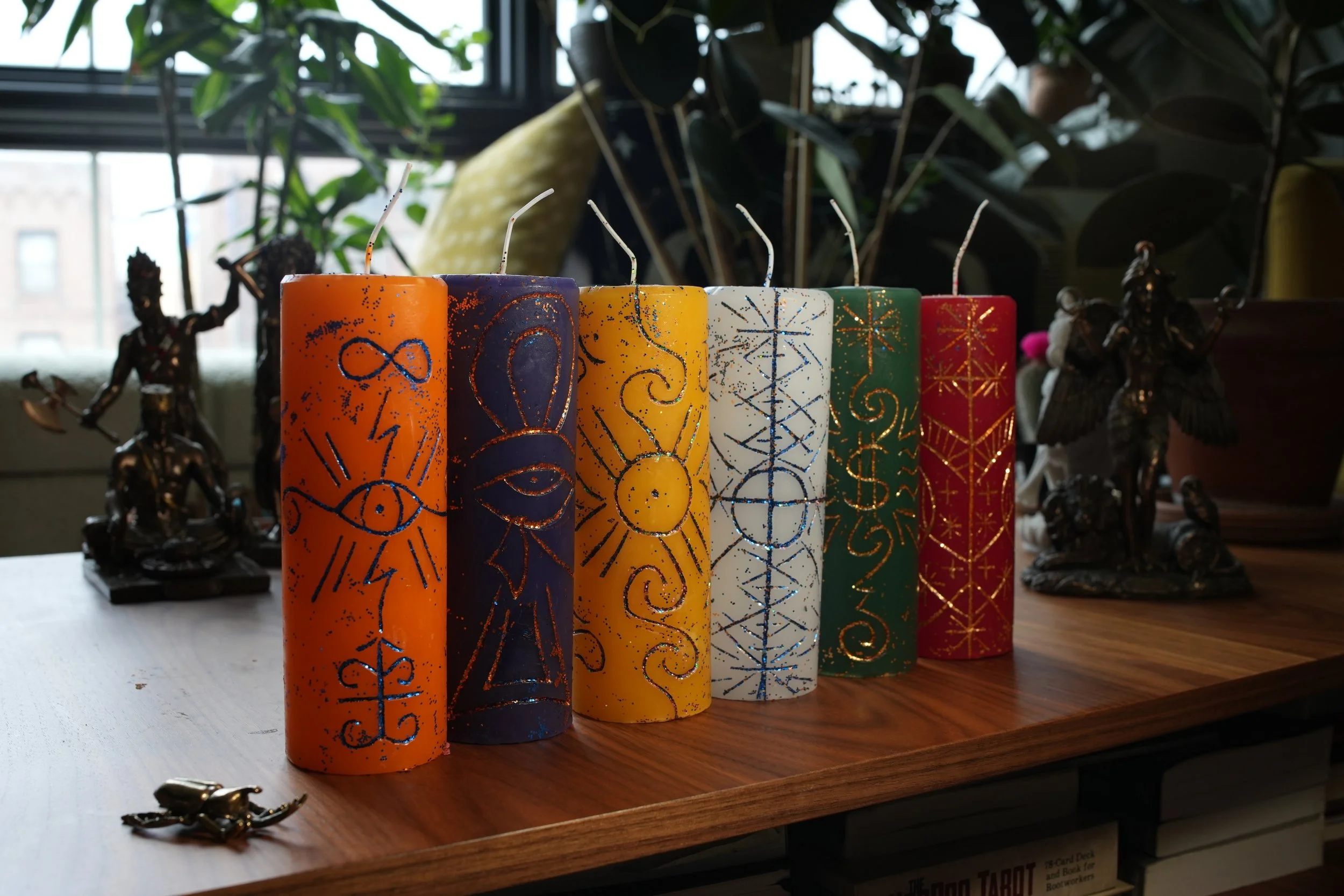 Seven colorful birthday candles arranged in a row on a wooden surface with various statues and plants in the background.