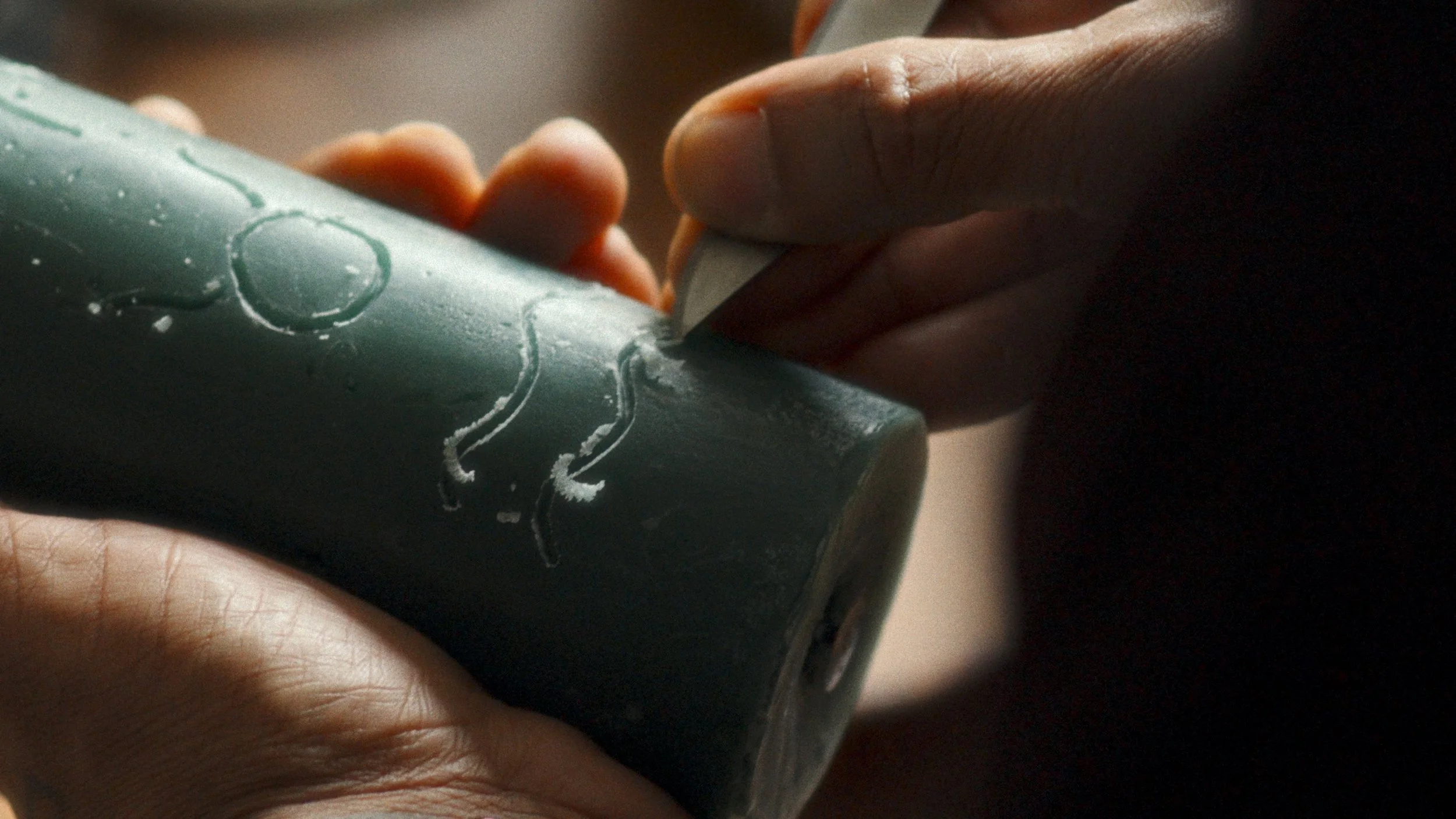 Close-up of a hand carving an intricate design sigil into a dark green candle, showing the detailed process of engraving.