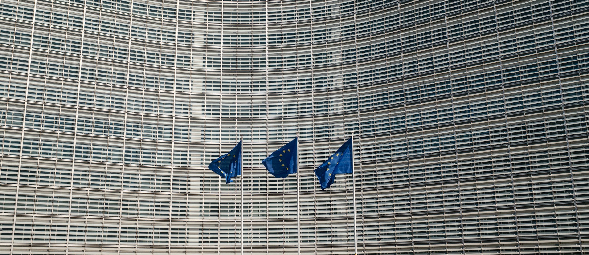 Three European Union flags flying in front of a modern building with a curved glass facade.