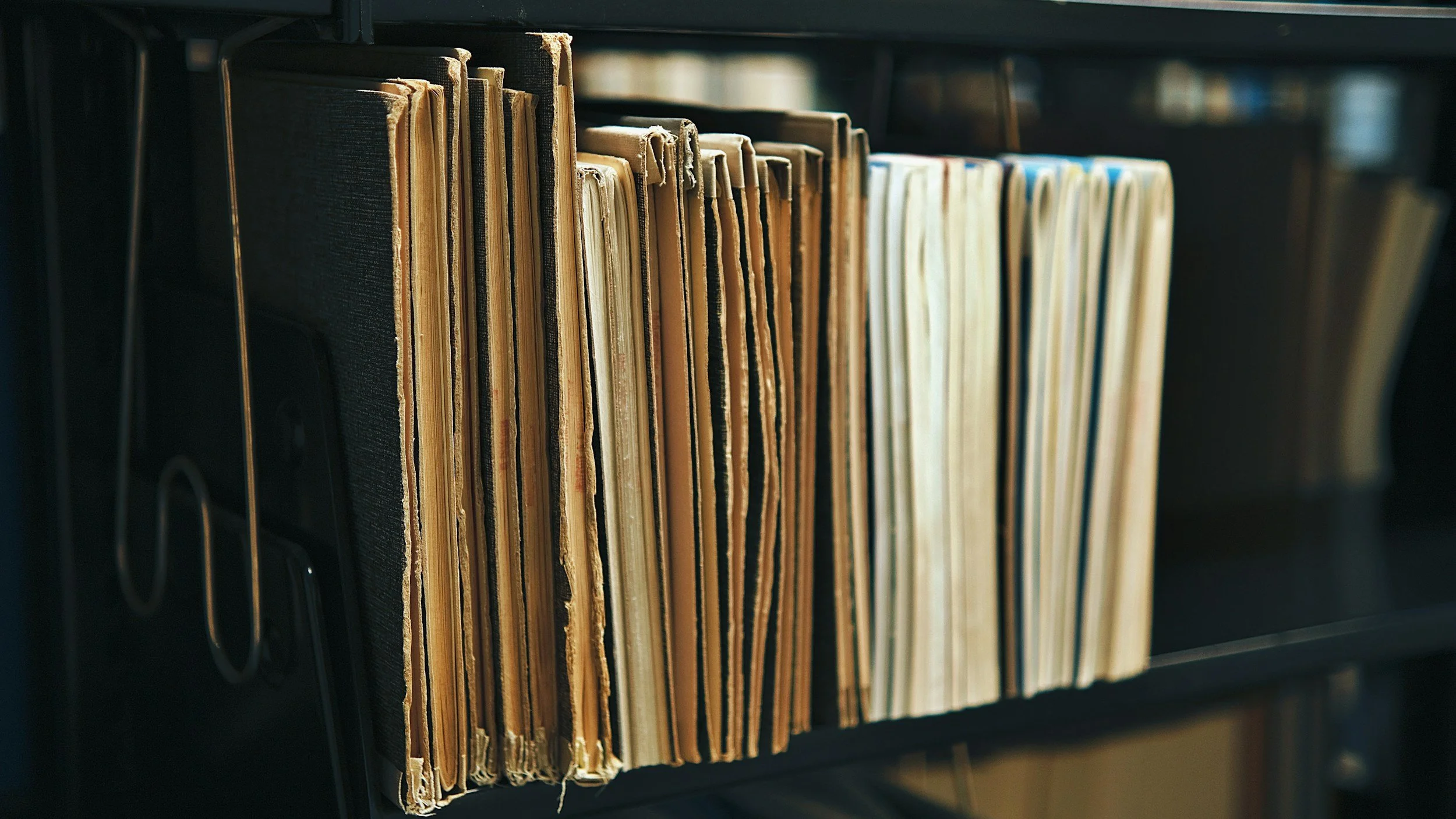 A row of worn, yellowed folders with torn edges on a metal shelf in a library or office setting.